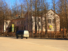 двухэтажный жилой дом с розовыми стенами вдоль дороги с желтыми дорожными знаками и движущимся транспортом в Сычево