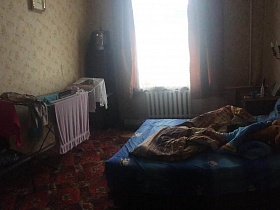 большая кровать с синим постельным, тумбочка, сушилка с бельем на красном ковре спальной комнаты с часами и бра, стилизованное под свечи на цветных стенах квартиры эпохи СССР