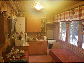 коричневая кухня, газовая плита с вытяжкой, микроволновка, электрочайчик и большой обеденный стол с клетчатой скатертью у окна с решеткой на кухне дачи эпохи СССР