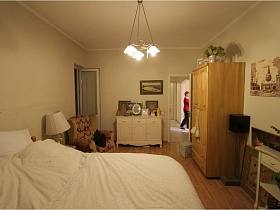 настольная лампа с белым плафоном на деревянной тумбочке у большой деревянной кровати с белым постельным,иконы, фотографии, зеркало на белом комоде, деревянный шкаф для одежды в светлой спальне простой семейной трехкомнатной квартиры