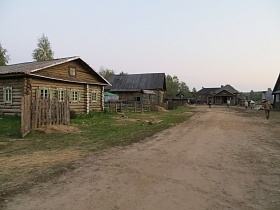 бревенчатые деревянные дома с невысоким забором вдоль широкой проселочной дороги в большой деревне начала 20-го века