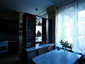 коричневые стулья со спинкой вокруг стола с мраморным покрытием у мебельной стенки, разделяющей кухню и гостиную квартиры художника