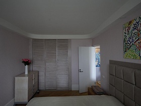 розовые розы в белой вазе на тумбе у трехдверного встроенного шкафа для одежды в спальной комнате стильной трешки минимализм в светлых тонах
