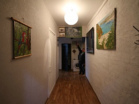 настенная вешалка для одежды, круглый плафон подвесной люстры на белом потолке длинного коридора с яркими картинами на стенах современной двухкомнатной квартиры