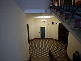вид на просторную лестничную площадку с белыми стенами, двухцветной плиткой на полу с жилыми квартирами и лифтом на этаже с высоты другого этажа