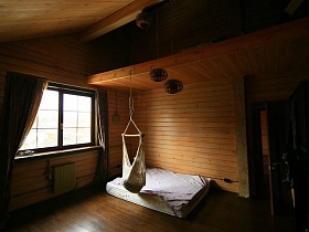 белый гамак у белого матраса на деревянном полу в углу комнаты с коричневыми шторами на большом окне в мансарде уютного дома в восемь комнат