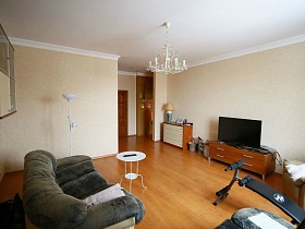 белый круглый столик у двухцветного углового дивана, навесные полки на светлой стене гостиной с комодом и телевизором на тумбе квартиры, удобной для переделок и ремонта в жилом доме