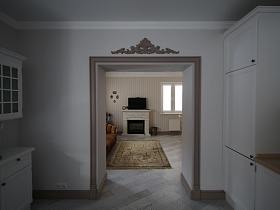 телевизор над белым камином, коричневый коврик у мягкого дивана  в гостиной из открытого дверного проема светлой кухни