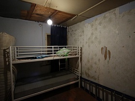 двухярусная металлическая кровать с матрасом у стены с поврежденными обоями и обычной лампочкой в разрушенном потолке небольшого домика отшельника у маленького озера