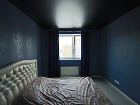 коричневое покрывало на большой белой кровати с рифленной мягкой спинкой у синей стены спальной комнаты с черным потолком и синими шторами на большом окне