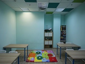 яркий детский коврик в стиле пэчворк на квадратном полу голубой классной комнаты с рядами учебных парт у голубых стен