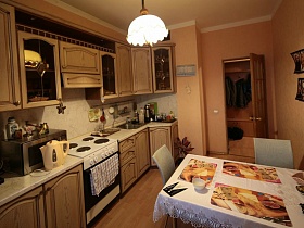 бежевая кухня с многочисленными шкафчиками, открытыми полочками, светлой мраморной столешницей в комнате с белой люстрой на потолке простой квартиры молодоженов