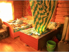 незаправленные кровати с подушками и постельным у стены с ковром, мягкое кресло у окна зонированной комнаты за цветной шторой в жилом доме в деревне