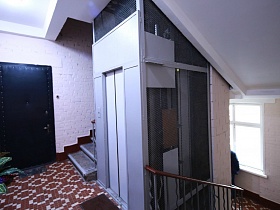 исскуственный цветок на полу двухцветной квадратной плитки на лестничной площадке с лестницей вокруг лифтовой кабины и ковриком у входной черной двери в квартиру художника