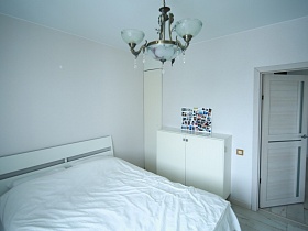 подвесная люстра с белыми стеклянными плафонами над белой кроватью с белым покрывалом в спальной комнате с открытой дверью в гостиную двухкомнатной квартиры