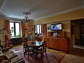 мягкие стулья со спинкой вокруг круглого стола на красном ковре в центре гостиной с ораньжевыми шторами на окне и в дверном проеме трехкомнатной квартиры времен СССР