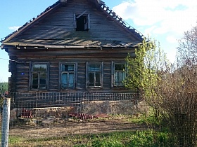 старый неокрашенный деревянный дом на высоком цоколе с открытым чердачным окном и полуразрушенной крышей за сетчатым забором