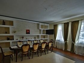 стулья со спинками вокруг длинного обеденного стола с салфетками у стены с полками из гипсокартона в столовой совмещенной с кухней современного кирпичного дома