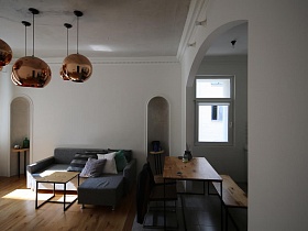 серый мягкий угловой диван с многочисленными подушками, деревянный журнальный столик на металлических квадратных ножках у белой стены с встроенными арочными нишами в гостиной зонированной комнаты современной скандинавской квартиры