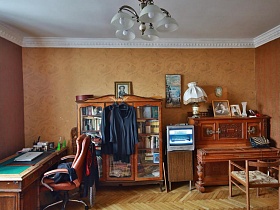 полукресло у пианино, книжный шкаф с книгами на полках, серебристый телевизор и длинный старинный письменный стол у стены персикого цвета в спальне трехкомнатной квартиры времен СССР