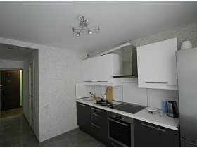 люстра в стиле хай-тек на белом потолке светлой кухни с белыми шкафами наверху, белой столешницей темного низа мебельной стенки однокомнатной квартиры