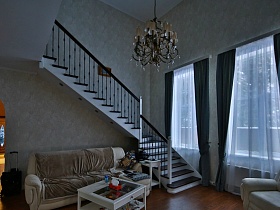 белый стеклянный столик у дивана с бежевым покрывалом под лестницей у окна светлой гостиной с темно серыми шторами кирпичного двухэтажного дома