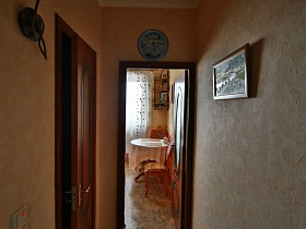 стулья со спинками вокруг круглого обеденного стола с белой скатертью на кухне через открытую дверь яркой трехкомнатной современной квартиры с комнатой бабушки