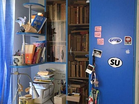 многочисленные книги на открытых полках углового компьютерного стола с белой столешницей и синими дверцами шкафа в спальной комнате квартиры в Бибирево