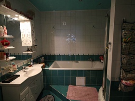 туалетные принадлежности на открытых полках у зеркала со шкафчиком над белой раковиной, белая ванна, зашитая зеленой плиткой на поддиуме бело-зеленой ванной комнаты современной трехкомнатной квартиры