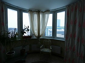 высокие комнатные цветы в вазонах, светлый мягкий стул со спинкой у эркерного окна с белой гардиной и цветными шторами просторной гостиной современной трехкомнатной квартиры