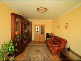 журнальны столик со стеклянной поверхностью у коричневого гладкого дивана, цветной ковер в центре гостиной трехкомнатной квартиры