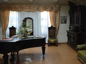 старинная мебель-черный рояль, шкаф из натурального дерева, кресла с высокими спинками, трюмо у окон с белой гардиной и бежевыми шторами школы-музея 19 века