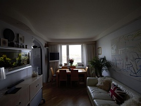 большой прямоугольный стол со стульями и белый телевизор у окна, шкаф и мягкий диван в просторной светлой гостиной современной трехкомнатной квартиры в Икеа стиле