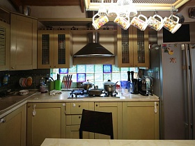 серебристый двухдверный холодильник у бежевой кухни с плитой и вытяжкой, лампы дневного света в подвесной люстре в зонированной комнате классической семейной дачи