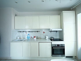 высокий белый холодильник у белой мебельной стенки с белой раковиной, белой вытяжкой над газовой поверхностью встроенной плиты, посуда на белой столешнице в зоне кухни просторной комнаты двухкомнатной квартиры