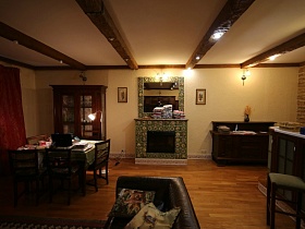 зеленая скатерть на обеденном столе, коричневый шкаф с посудой за стеклянными дверцами и коричневый комод по обе стороны декоративного камина с зеленой плиткой и зеркалом на стене просторной кухни с островком готовки
