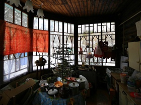газовая плита, шкафы, круглый стол по центру с клеенкой и многочисленной посудой и исскуственной елочкой на веранде деревянной дачи художника с овальной террасой времен СССР