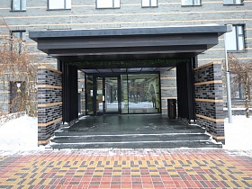 плитка в серых тонах на боковых стенах просторного крыльца со ступенями под навесом со стеклянными входными дверьми в подъезд современного жилого дома
