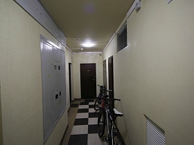 велосипеды на шахматном полу светлого длинного коридора с электрическим щитом, вентиляционными решетками и дверьми в квартиры этажа современного многоэтажного дома