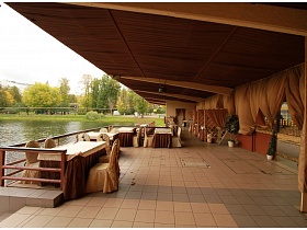 уютные столики со стульями у деревянных перил открытого кафе на озере с большим навесом над просторной площадкой