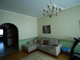 светло серый мягкий угловой диван у стены с салатовыми панельными обоями светлой гостиной с большой люстрой на белом потолке простой семейной квартиры на втором этаже в Марьино