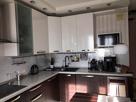 белые верхние шкафы темной мебельной стенки с кухонными электроприборами на светлой столешнице, телевизор на полке в кухне с полосатыми бежевыми обоями белым потолком