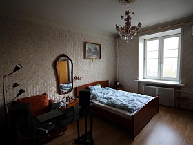 черный торшер в стиле хай-тек, деревянные часы на прикроватной тумбочке в углу, табурет у окна в спальной комнате квартиры сталинки