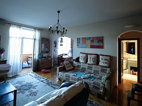 люстра, стилизованная под свечи над большим голубым ковром в гостиной с открытой дверью на балкон в семейной трешке