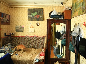 рыжая кошка на деревянной кровати с бежевым покрывалом у стены с простым ковриком и картинами,коричневый шкаф с зеркальной дверцей в спальне советской дачи художника с овальной террасой