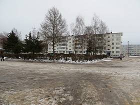 белый пятиэтажный жилой дом за сквером с раскидистыми елями , кустарниками и лиственными деревьями у здания  ДК СССР