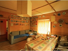 общий вид просторной гостиной с цветной скатертью на обеденном столе, синим покрывалом на сером мягком угловом диване у стены с разнообразными декоративными тарелками