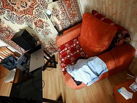 яркое ораньжевое кресло с подушкой под коричневым цветным покрывалом, черный стеклянный столик у стены с цветным ковром в спальной комнате квартиры сталинки