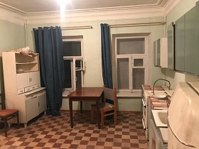 белый кухонный шкаф, обеденный стол у окон с белыми деревянными рамами и синими шторами на кухне с серо-зеленой мебельной стенкой на полу с коричневой шахматной плиткой