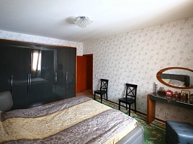 овальное зеркало над прямоугольным столиком с мягким пуфиком, черные деревянные стулья со спинкой у стены с цветочными светлыми обоями спальной комнаты с люстрой на белом потолке